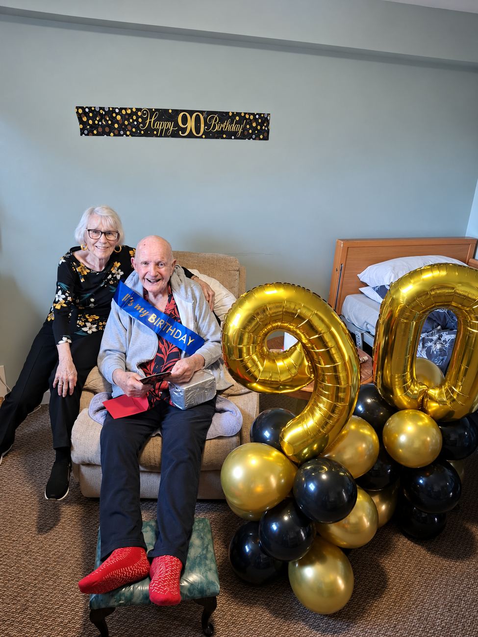 A resident at Higher Park Lodge celebrating his 90th birthday, with gold balloons and a family member beside him.