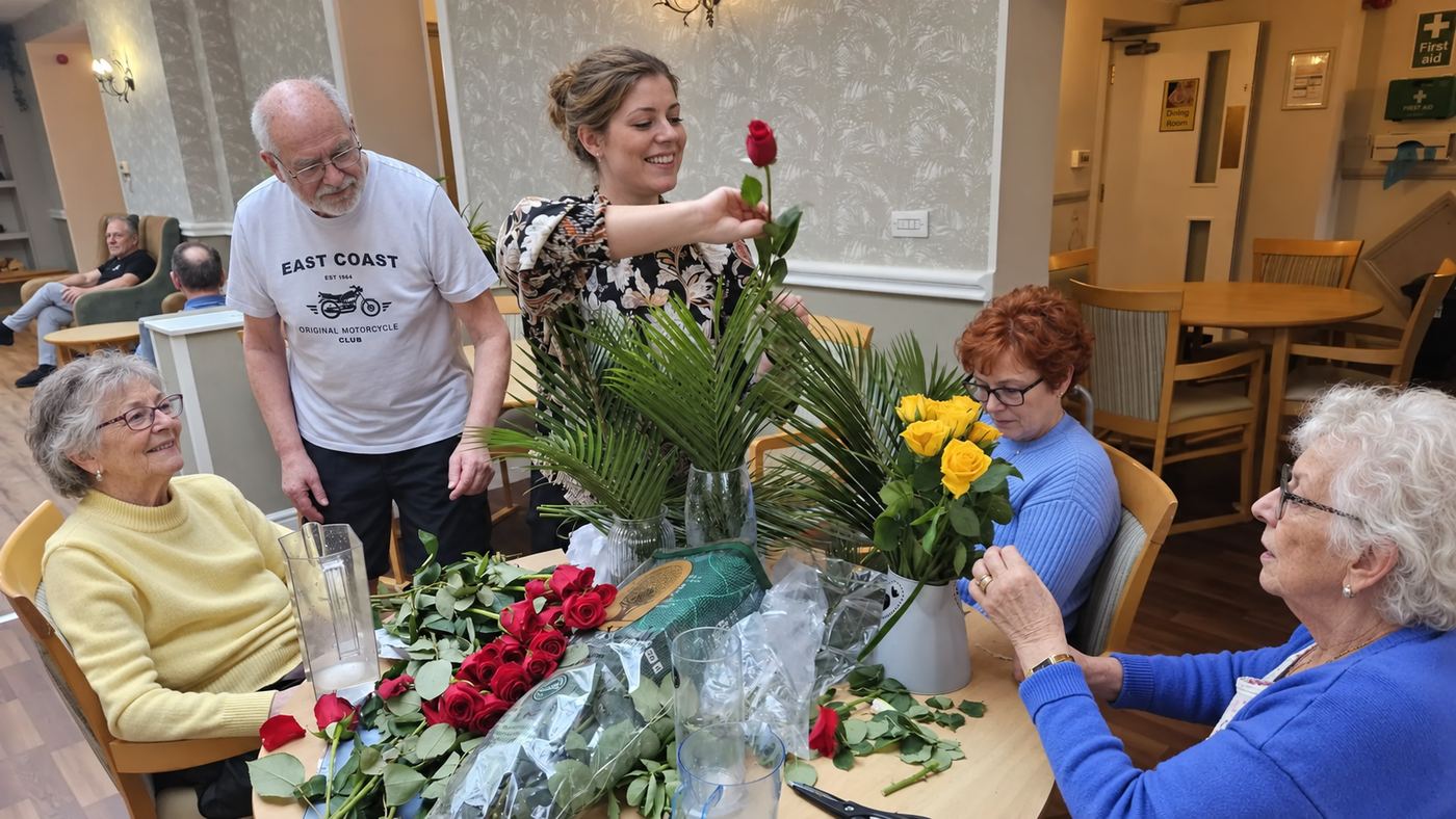 A carer arranging flowers with a group of residents around a table.