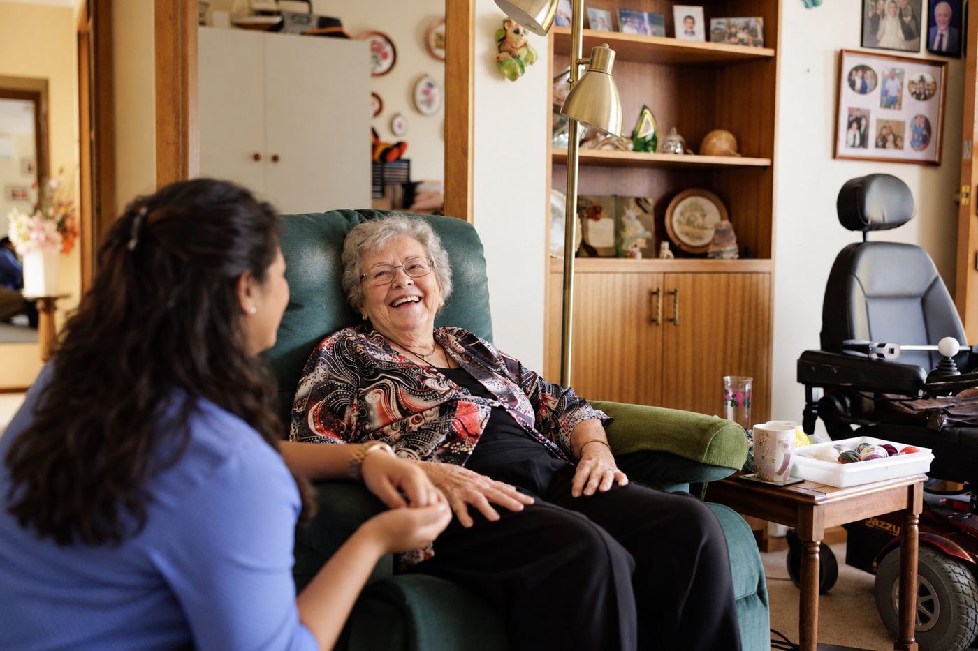 A resident laughing with a carer in her room, sitting in her armchair among her own pictures and things.